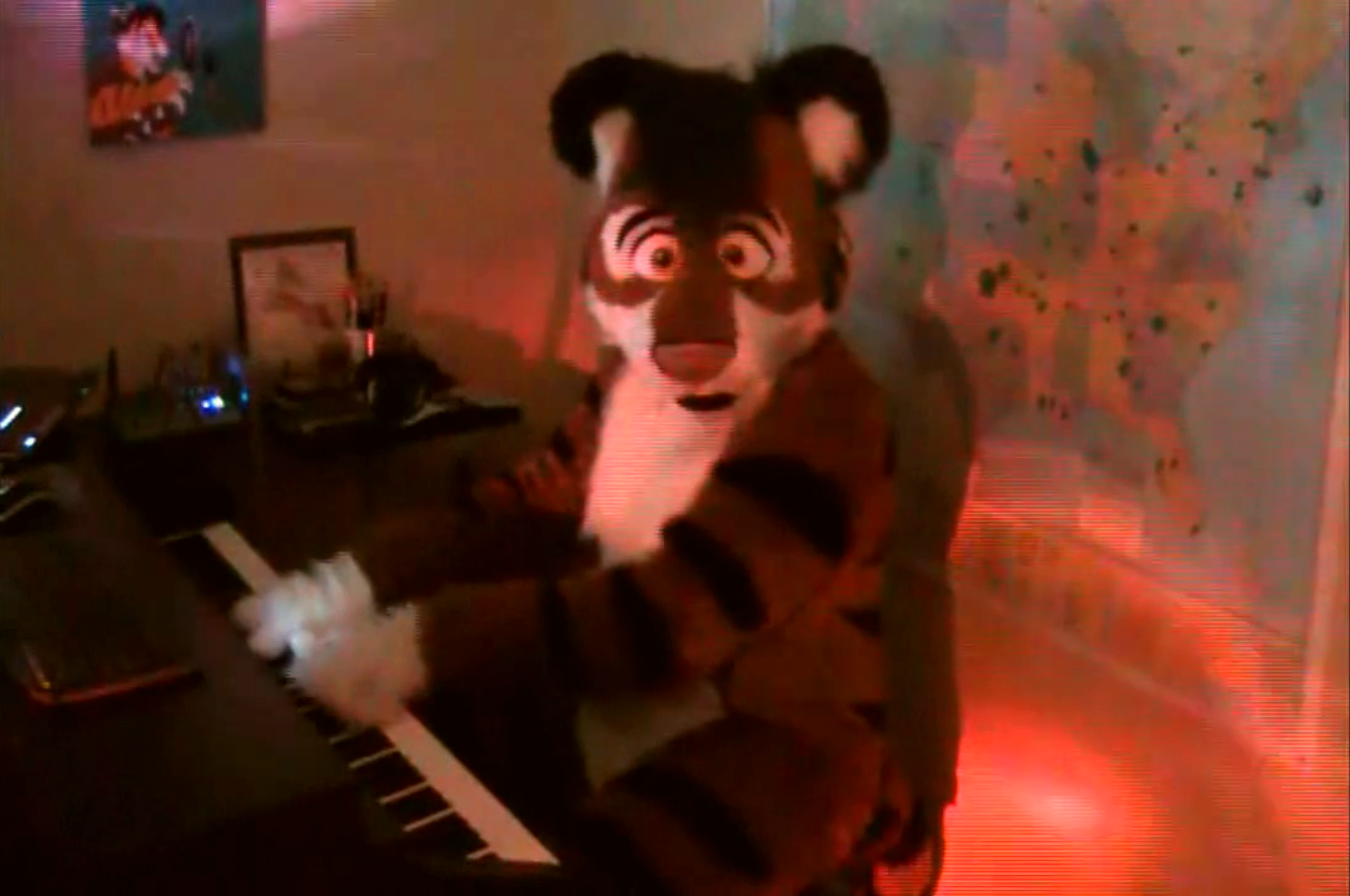 A man in a fursuit is playing piano while looking at the viewer through the camera. The fursuit is that of a tiger. Inside the room there is a map, piano, router, and various placards and paintings.