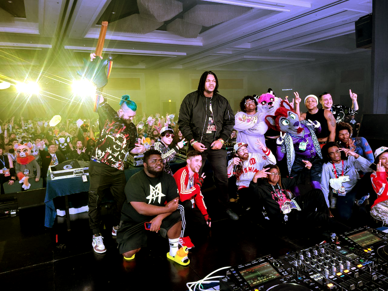 In the foreground of the image is a collection of various people of whom some are in fursuits, posing either by kneeling or standing and holding a 'peace' sign with two fingers in the air. A DJ deck is visible in the corner. To the back is an audience full of fursuiters and unsuited audience members similarly doing 'peace signs'.