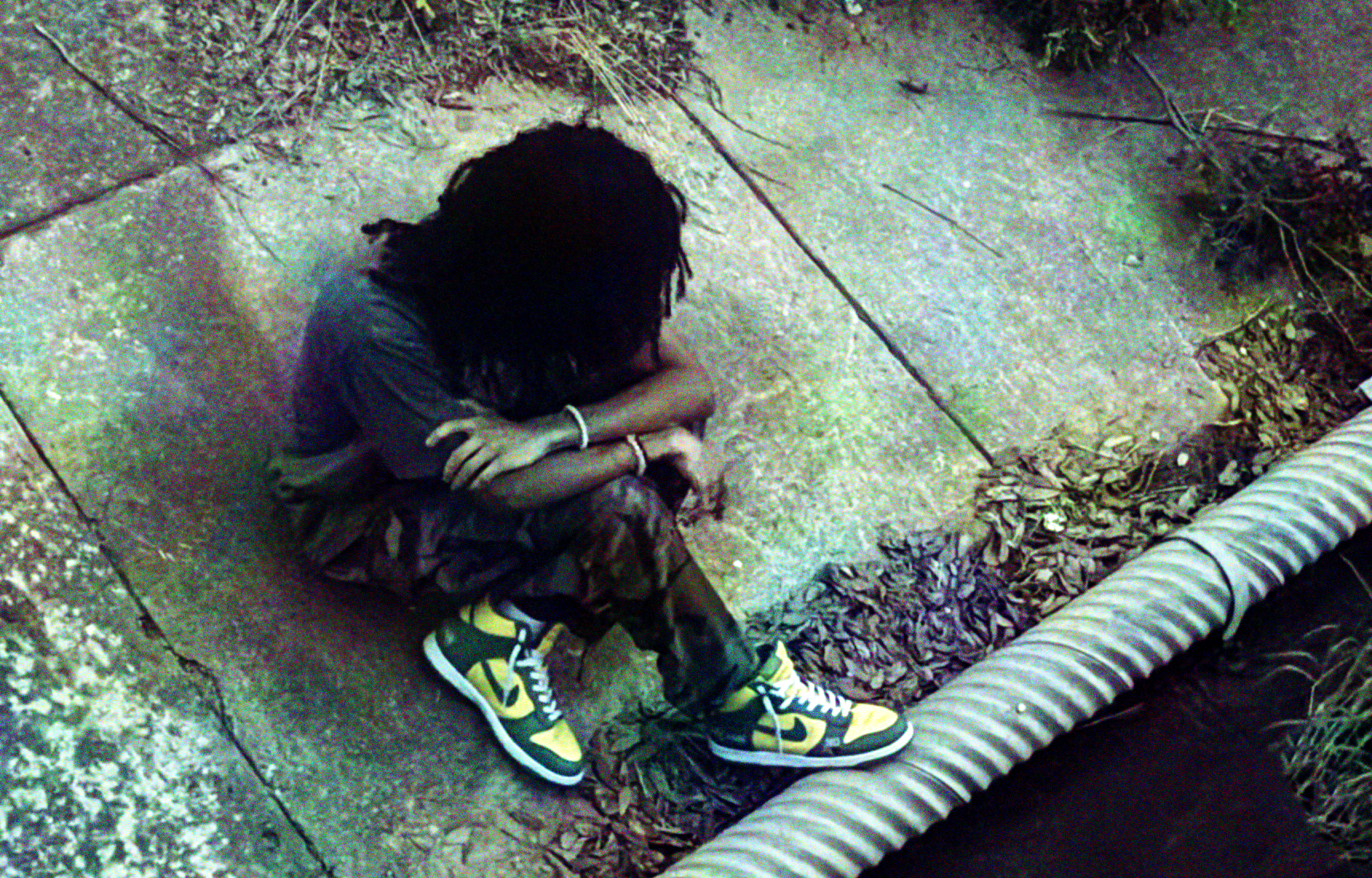 A woman with dreads, two bracelets around each arm, and Nike sneakers sits on the sidewalk with arms folded. There is a bunch of foliage around. Her right foot is grazing what looks to be a pipe.